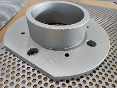 A metallic circular component with multiple holes is placed on a perforated surface. The component has a cylindrical section protruding from a flat base.