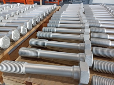A neatly arranged collection of large metal bolts with hexagonal heads and threaded bodies, aligned in multiple rows on a wooden pallet, suggesting an orderly industrial setting.