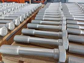 A neatly arranged collection of large metal bolts with hexagonal heads and threaded bodies, aligned in multiple rows on a wooden pallet, suggesting an orderly industrial setting.