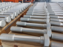 A neatly arranged collection of large metal bolts with hexagonal heads and threaded bodies, aligned in multiple rows on a wooden pallet, suggesting an orderly industrial setting.