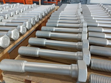 A neatly arranged collection of large metal bolts with hexagonal heads and threaded bodies, aligned in multiple rows on a wooden pallet, suggesting an orderly industrial setting.