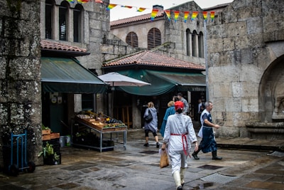 A bustling outdoor marketplace is set against the backdrop of historic stone buildings. Several people walk through the area, some dressed in traditional costumes. A fruit and vegetable stand is visible under a green awning, with produce neatly arranged. Colorful triangular banners are strung overhead, adding vibrancy to the scene.