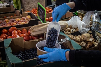 A market scene with a variety of fresh produce including tomatoes, peaches, and mushrooms displayed in open boxes. A person wearing blue gloves is scooping blueberries from a carton into a bowl, with a focus on the action. A price sign for tomatoes is visible in the background.