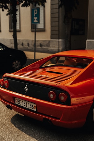 A bright red Ferrari is parked on a street, showcasing its sleek design and iconic rear lights. The car is next to a parking sign and surrounded by buildings.