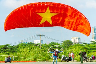 A person engaged in paragliding with a large red parachute featuring a yellow star in the center, reminiscent of the Vietnam flag. The individual wears a helmet and gear, preparing for takeoff in a spacious, outdoor area with green foliage and urban buildings visible in the background. Bystanders observe the scene.