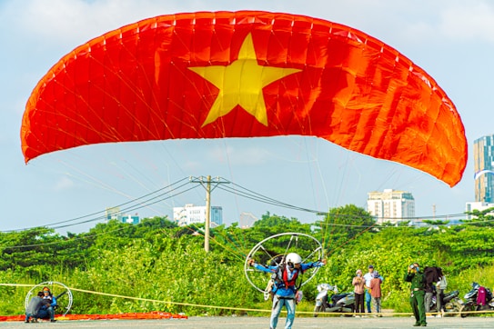 A person engaged in paragliding with a large red parachute featuring a yellow star in the center, reminiscent of the Vietnam flag. The individual wears a helmet and gear, preparing for takeoff in a spacious, outdoor area with green foliage and urban buildings visible in the background. Bystanders observe the scene.