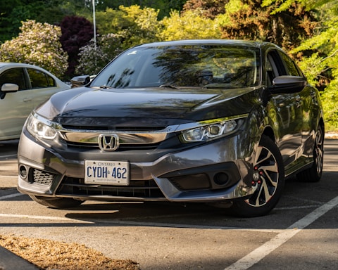 A dark gray Honda sedan is parked in a shaded outdoor parking lot with a visible Ontario license plate. The scene is set in a landscaped area with lush greenery and a few other cars in the background. Sunlight creates reflections on the car's surface, highlighting its sleek design.