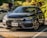 A dark gray Honda sedan is parked in a shaded outdoor parking lot with a visible Ontario license plate. The scene is set in a landscaped area with lush greenery and a few other cars in the background. Sunlight creates reflections on the car's surface, highlighting its sleek design.