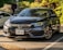 A dark gray Honda sedan is parked in a shaded outdoor parking lot with a visible Ontario license plate. The scene is set in a landscaped area with lush greenery and a few other cars in the background. Sunlight creates reflections on the car's surface, highlighting its sleek design.