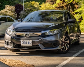 A dark gray Honda sedan is parked in a shaded outdoor parking lot with a visible Ontario license plate. The scene is set in a landscaped area with lush greenery and a few other cars in the background. Sunlight creates reflections on the car's surface, highlighting its sleek design.
