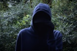 A figure wearing a dark hoodie stands in front of dense greenery, partially obscuring their face with one hand. The background is filled with lush green leaves, creating an atmosphere of mystery and concealment.