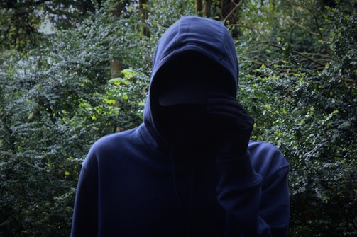 A figure wearing a dark hoodie stands in front of dense greenery, partially obscuring their face with one hand. The background is filled with lush green leaves, creating an atmosphere of mystery and concealment.