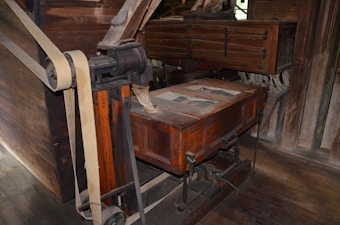 A vintage wooden and metal machine with intricate parts, including large belts and wheels, likely used for industrial or milling purposes. The structure appears aged, with visible wear on the wood and metal components.