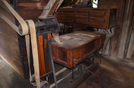 A vintage wooden and metal machine with intricate parts, including large belts and wheels, likely used for industrial or milling purposes. The structure appears aged, with visible wear on the wood and metal components.