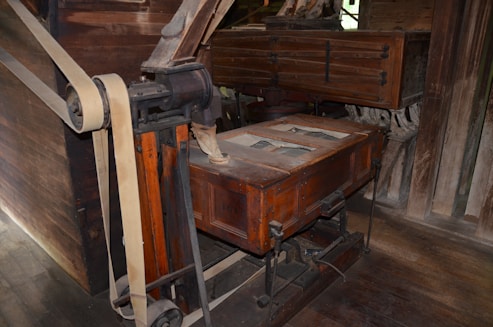 A vintage wooden and metal machine with intricate parts, including large belts and wheels, likely used for industrial or milling purposes. The structure appears aged, with visible wear on the wood and metal components.