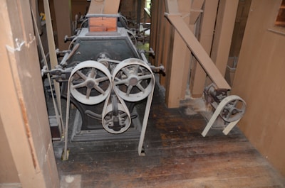 An old industrial machine with large metal gears and pulleys is positioned on a wooden floor. The machine's components appear worn and rusty, surrounded by a wooden structure with beams and panels.