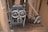 An old industrial machine with large metal gears and pulleys is positioned on a wooden floor. The machine's components appear worn and rusty, surrounded by a wooden structure with beams and panels.