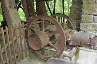 A large, old, rusted industrial machine with metal gears and wheels, surrounded by wooden and stone elements, is set in an outdoor environment with greenery visible in the background.