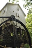 A rustic wooden watermill with a large black metal water wheel located beside a stone base. The mill structure is surrounded by lush greenery, with trees providing shade. The building has multiple windows, some with open shutters, and the overall scene suggests an old-fashioned, tranquil setting.