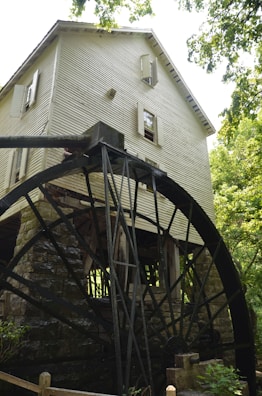 A rustic wooden watermill with a large black metal water wheel located beside a stone base. The mill structure is surrounded by lush greenery, with trees providing shade. The building has multiple windows, some with open shutters, and the overall scene suggests an old-fashioned, tranquil setting.