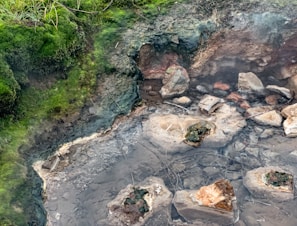A natural hot spring surrounded by lush green moss and rocky terrain. The steam rises from the water, indicating its warmth. The water is clear, revealing rocks of various shapes and sizes submerged. The edges are lined with vibrant green moss and some patches of grass.