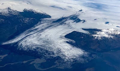 A vast, icy glacier stretches across a rugged terrain, with intricate patterns of white and dark lines creating a mesmerizing view. Snow-capped mountains flank the glacier, extending into what appears to be a cloudy sky.