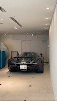 A sleek, black sports car is parked indoors on a light-colored tiled floor. The vehicle has a futuristic design with a glossy finish and visible aerodynamic features such as a rear wing and diffuser. It is positioned against a neutral-colored wall with minimalistic decor.
