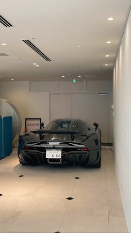 A sleek, black sports car is parked indoors on a light-colored tiled floor. The vehicle has a futuristic design with a glossy finish and visible aerodynamic features such as a rear wing and diffuser. It is positioned against a neutral-colored wall with minimalistic decor.