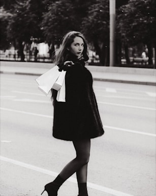 A stylish woman walks down a city street in a black fur coat and high-heeled boots, holding shopping bags. The scene is in black and white, lending it a timeless, classic feel with trees and a road as the background.