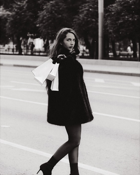 A stylish woman walks down a city street in a black fur coat and high-heeled boots, holding shopping bags. The scene is in black and white, lending it a timeless, classic feel with trees and a road as the background.