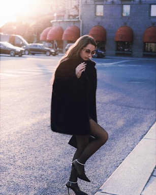 A person wearing a fur coat and sunglasses walks along an urban street. The setting sun casts a warm glow, and red awnings line the building in the background. The scene captures a moment of city life with a touch of elegance.