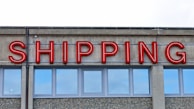 A concrete building facade displays the word 'SHIPPING' in bold, red neon letters above a series of uniform, rectangular windows with reflective glass.