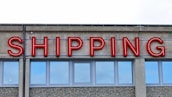 A concrete building facade displays the word 'SHIPPING' in bold, red neon letters above a series of uniform, rectangular windows with reflective glass.