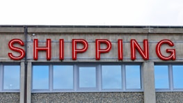 A concrete building facade displays the word 'SHIPPING' in bold, red neon letters above a series of uniform, rectangular windows with reflective glass.