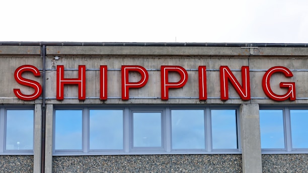 A concrete building facade displays the word 'SHIPPING' in bold, red neon letters above a series of uniform, rectangular windows with reflective glass.