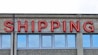 A concrete building facade displays the word 'SHIPPING' in bold, red neon letters above a series of uniform, rectangular windows with reflective glass.
