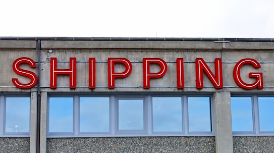A concrete building facade displays the word 'SHIPPING' in bold, red neon letters above a series of uniform, rectangular windows with reflective glass.
