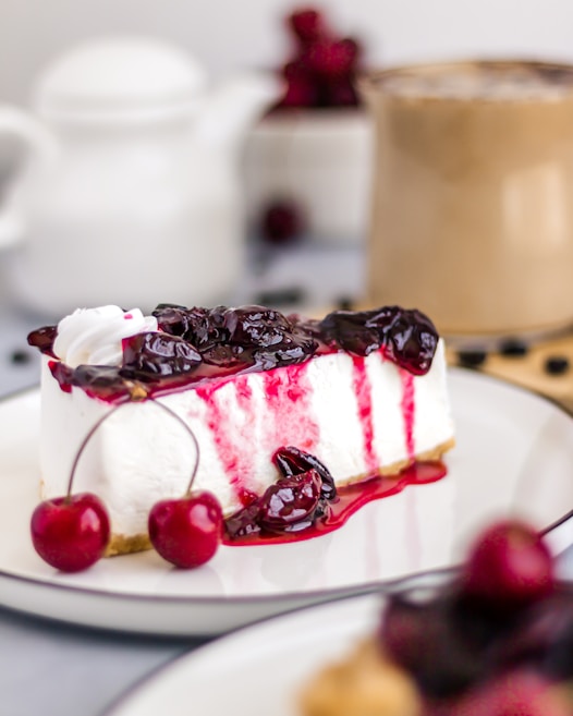 A close-up of a slice of cheesecake topped with a generous amount of cherry compote, with whole cherries on the side. The cheesecake is presented on a white plate, with a blurred background that includes a cup of coffee and what appears to be a teapot.