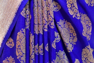 A detailed view of a richly embroidered textile with a vibrant blue background. Intricate golden floral and geometric patterns adorn the fabric, creating a luxurious and traditional appearance. The edge of the textile features a subtle herringbone design.