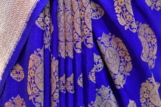 A detailed view of a richly embroidered textile with a vibrant blue background. Intricate golden floral and geometric patterns adorn the fabric, creating a luxurious and traditional appearance. The edge of the textile features a subtle herringbone design.