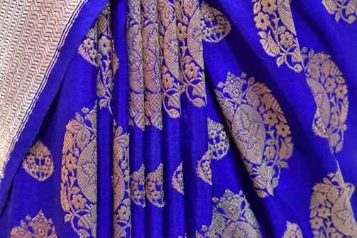 A detailed view of a richly embroidered textile with a vibrant blue background. Intricate golden floral and geometric patterns adorn the fabric, creating a luxurious and traditional appearance. The edge of the textile features a subtle herringbone design.