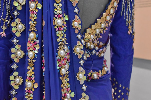 A close-up view of an ornately decorated fabric featuring large pearls, golden floral patterns, and colorful gemstones in a rich blue material. The fabric appears luxurious and intricate, with detailed embroidery and embellishments forming floral and geometric shapes.