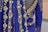 A close-up view of an ornately decorated fabric featuring large pearls, golden floral patterns, and colorful gemstones in a rich blue material. The fabric appears luxurious and intricate, with detailed embroidery and embellishments forming floral and geometric shapes.