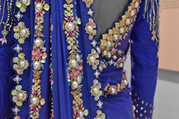 A close-up view of an ornately decorated fabric featuring large pearls, golden floral patterns, and colorful gemstones in a rich blue material. The fabric appears luxurious and intricate, with detailed embroidery and embellishments forming floral and geometric shapes.