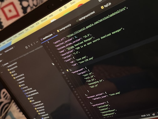 A computer screen displays a code editor with a dark theme. The code is related to a manifest file, showing JSON format data, including URLs, version numbers, and a description of an 'Xtreme Download Manager'. There are also multiple tabs open with filenames like 'background.html' and 'bg2.js'.