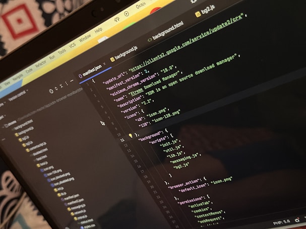 A computer screen displays a code editor with a dark theme. The code is related to a manifest file, showing JSON format data, including URLs, version numbers, and a description of an 'Xtreme Download Manager'. There are also multiple tabs open with filenames like 'background.html' and 'bg2.js'.