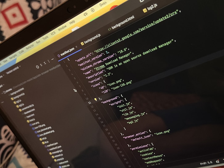 A computer screen displays a code editor with a dark theme. The code is related to a manifest file, showing JSON format data, including URLs, version numbers, and a description of an 'Xtreme Download Manager'. There are also multiple tabs open with filenames like 'background.html' and 'bg2.js'.