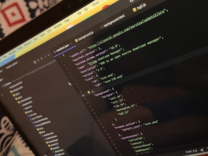 A computer screen displays a code editor with a dark theme. The code is related to a manifest file, showing JSON format data, including URLs, version numbers, and a description of an 'Xtreme Download Manager'. There are also multiple tabs open with filenames like 'background.html' and 'bg2.js'.