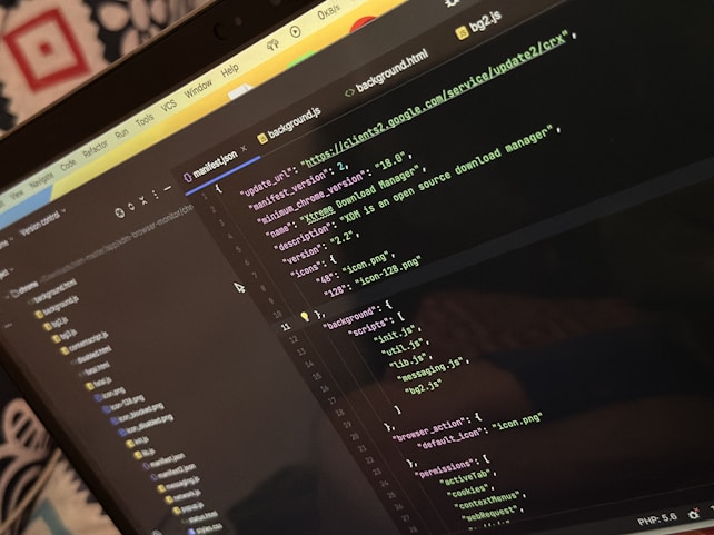 A computer screen displays a code editor with a dark theme. The code is related to a manifest file, showing JSON format data, including URLs, version numbers, and a description of an 'Xtreme Download Manager'. There are also multiple tabs open with filenames like 'background.html' and 'bg2.js'.