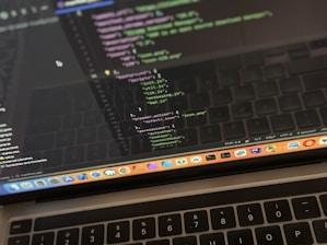 A close-up view of a laptop displaying code on the screen. The code editor is open on a dark-themed interface, showing lines of JavaScript with syntax highlighting. Below the screen, the keyboard of the laptop is partially visible. The laptop's dock at the bottom houses several application icons.
